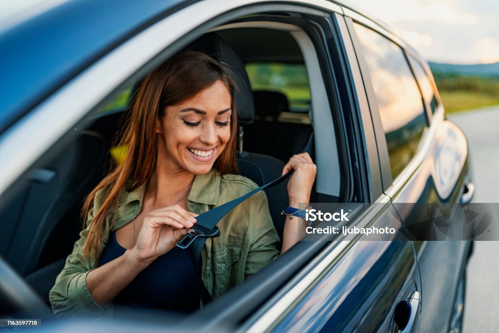 Photo of a business woman sitting in a car putting on her seat belt Young attractive woman sitting on car seat and fastening seat belt, car safety concept. Photo of beautiful young woman fastens a seat belt in the car - outdoors. Brown hair woman fastening seat belt in the car, safety concept. Business lady, Caucasian woman driver fastening car seat belt while sitting behind the wheel car. Safe driving concept. Selective focus, copy space Car Stock Photo