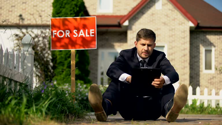 Tired estate broker sitting on ground near for sale signboard, failed contract