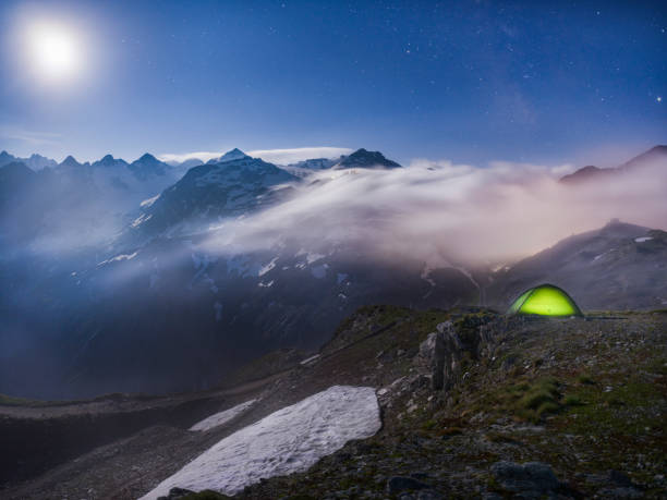istock Moonlit scene with illuminated green tent under starry night - fog rolling over mountains 1163614364