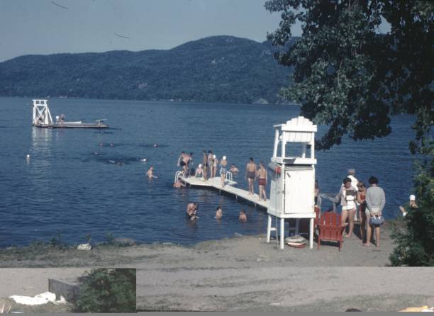 United States - January 01, 1965:  Vintage, authentic archival photograph of people lining up and diving off a swimming pier on a small beach at a lake outdoors, 1965