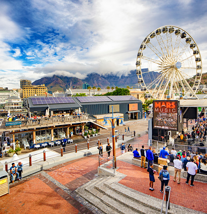 Cape Town Victoria and Albert waterfront with Ferris wheel and commercial pier. on this elevated view, people are attending an outdoors entertainment event or eating out on a restaurant patio. Table mountain in the background is partly covered by clouds.