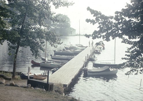 United States - January 01, 1965:  Vintage, authentic archival photograph of a pier leading into a lake among trees with boats and small sailboats moored at the pier, 1965