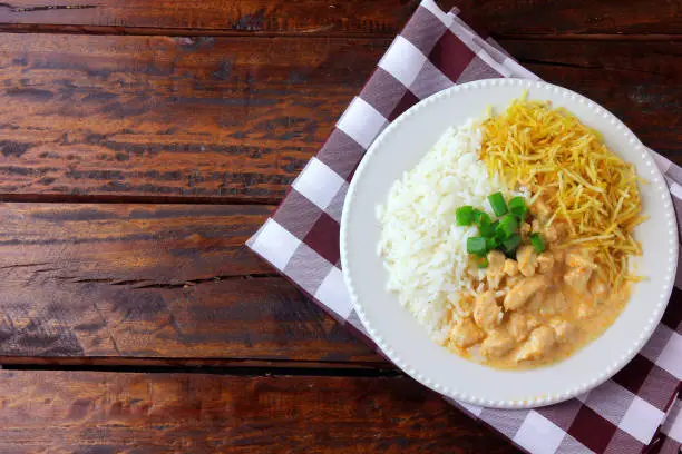 Photo of Chicken stroganoff, is a dish originating from Russian cuisine that in Brazil is composed of sour cream with tomato extract, rice and potato chips, on rustic wooden table. Top view.