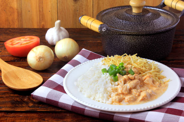 Chicken stroganoff, pan and ingredients. In Brazil it is composed of sour cream with tomato extract, rice and potato sticks, on rustic wooden table. Originating in Russian Cuisine stock photo