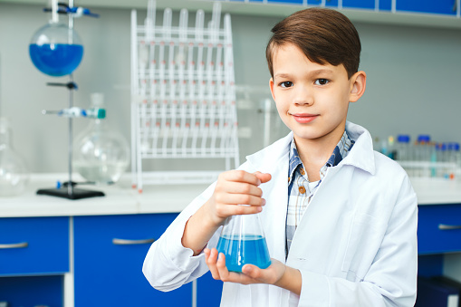 Little boy learning in school laboratory standing looking camera smiling holding test-tube with blue liquid for experiment