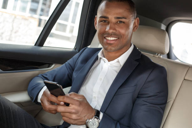 istock Young businessman sitting on back seat in car using smartphone looking camera happy close-up 1163630908