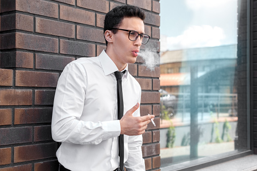 Young business man wearing eyeglasses standing leaning on wall on the city street holding cigarette smoking relaxed