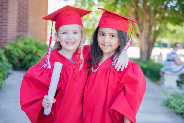 Beautiful elementary age girls wearing graduation caps and gowns smile while standing outside school after graduation ceremony stock photo
