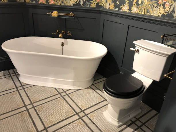 Stock Photo of luxury washroom bathroom with large freestanding bath with curved ends and bronze mixer tap, white old fashioned toilet WC with wooden lid, wooden panelling painted black and floral wallpaper with flowers pattern, mosaic tiled flooring tiles imagen de baño de baño de lujo con gran baño independiente con extremos curvos y grifo mezclador de bronce, wc wc antiguo blanco con tapa de madera, paneles de madera pintados de negro y papel pintado floral con patrón de flores, baldosas de mosaico de