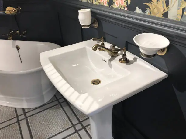 Stock Photo of luxury washroom bathroom with large freestanding bath with curved ends and bronze mixer tap, white rectangular sink wash basin with soap dish, wood panelling painted black and floral wallpaper with flowers pattern, mosaic tiled flooring tiles
