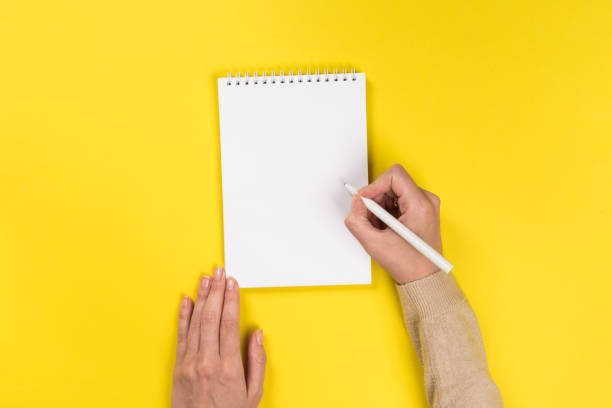 istock Woman's hands with perfect manicure holding pencil and  notepad as mockup for your design. Yellow background, flat lay 1163665076