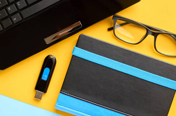 istock Black glasses, notebook, usb flash and laptop keyboard on blue and yellow background composition. 1163665170