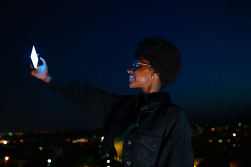 Cool young black woman taking a selfie with the city lights at background.