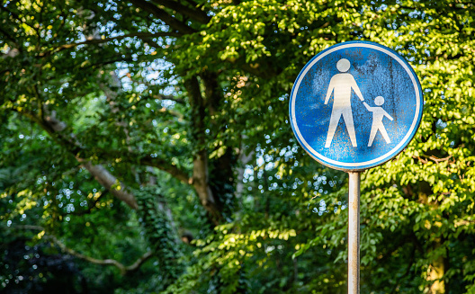 Attention, children crossing. White mother holding a child icon on round blue color sign, green trees outdoors background. Dutch road sign, Rotterdam Netherlands