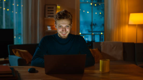 istock Handsome Smiling Man Works on a Laptop while Sitting at His Desk at Home. Young Freelancer Works on Computer in His Cozy Living Room with Warm Evening Lighting Turned on. 1163674337