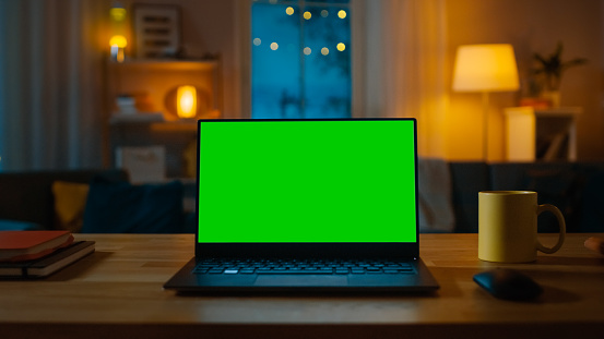 Laptop Computer Showing Green Chroma Key Screen Stands on a Desk in the Living Room. In the Background Cozy Living Room in the Evening with Warm Lights on.