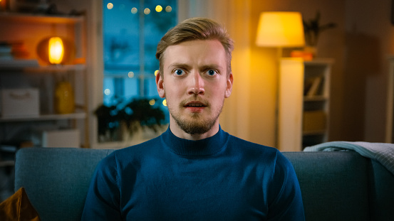 Portrait of a Handsome Man Sitting on a Couch at Home at Night, Watching Gore Horror Movie on TV. He Gets Really Scared, Confused and Disgusted.