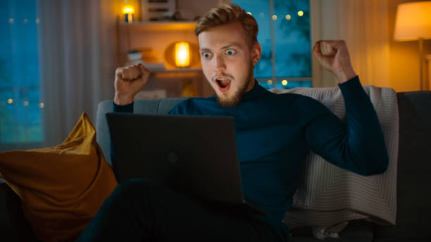 istock In the Evening: Portrait of Handsome Young Man Using Laptop while Sitting on a Couch at Home, Suddenly He Wins Big and Celebrates His Victory with YES Gesture. 1163674573