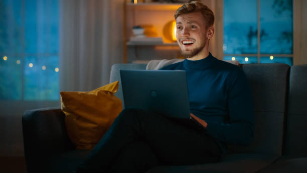istock In the Evening Handsome Young Man at Home Sitting on a Couch Works on a Laptop Computer and Watches Television Comedy and Laughs. 1163674591