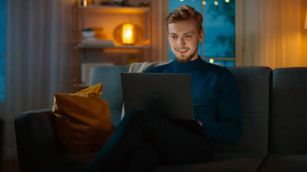 istock In the Evening Handsome Young Man at Home Sitting on a Couch Works on a Laptop Computer. In the Background Cozy Living Room. 1163674634