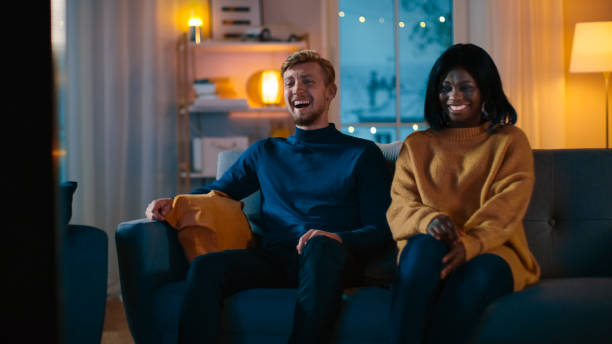 istock Happy Diverse Young Couple Watching Comedy on TV while Sitting on a Couch, they Laugh and Enjoy Show. Handsome Caucasian Boy and Black Girl in Love Spending Time Together in the Cozy Apartment. 1163674680