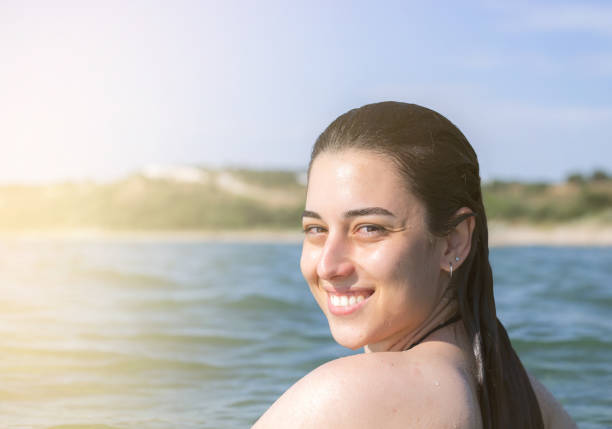 A young girl with short blond hair smiles brightly, standing near a river on a sunny day. Sunlight reflects off the water, creating a cheerful atmosphere around her.