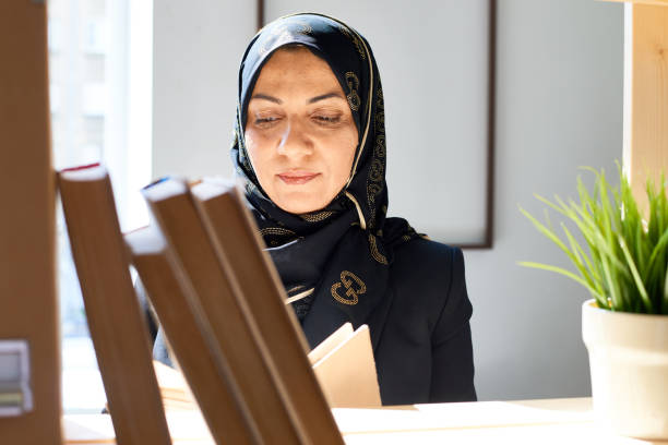 Ethnic woman in hijab with book Adult Muslim woman reading book standing at wooden shelf in sunlight middle eastern ethnicity mature adult book reading stock pictures, royalty-free photos & images