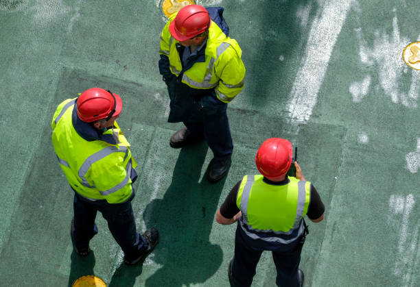 View from top deck of passenger ferry, looking at three ferry workers standing below 12th June 2019: Leaving Oban harbour on the northwest coast of Scotland. Looking down from the upper deck to the parking deck of the roll on roll off car & passenger ferry, where three crew workers in bright high visibility jackets & red hard hats are standing together, preparing to guide car drivers on board.  One of the men is talking into a walk-talkie handheld radio. oban stock pictures, royalty-free photos & images