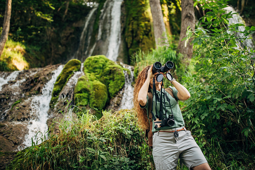 One woman, lady explorer and biologist standing in nature by waterfall alone, using binoculars.