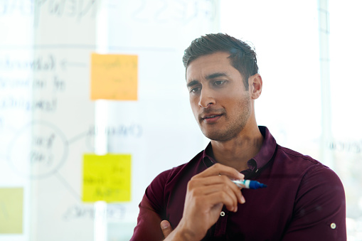 Cropped shot of a handsome young businessman looking thoughtful while writing notes on a glass wall in the office