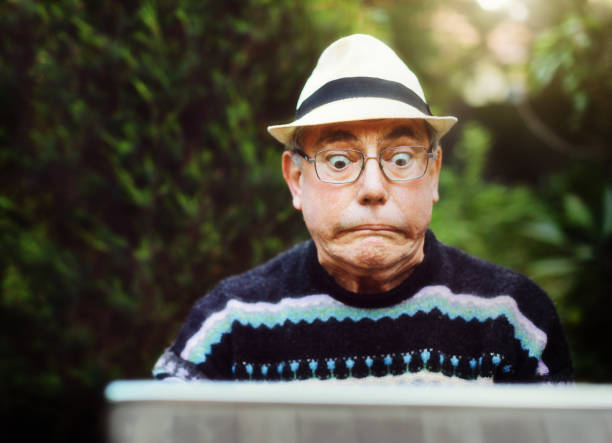 Senior man using laptop outdoors is confused and frustrated A senior man sitting in a garden using his laptop has a problem and glares at it, pulling a humorous face. shocked computer stock pictures, royalty-free photos & images
