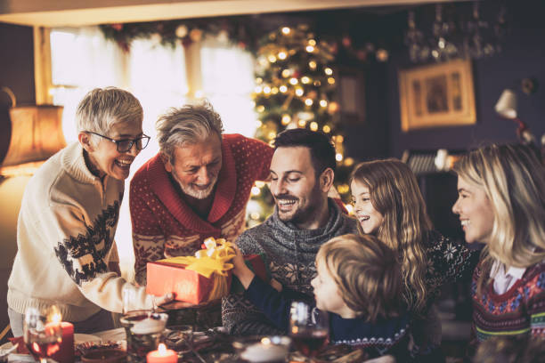 Time for Christmas presents! Happy senior grandparents giving Christmas presents to their family in dining room. multi generation family christmas stock pictures, royalty-free photos & images