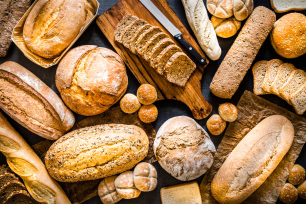 Breads assortment background Top view of various kinds of breads like brunch bread, rolls, wheat bread, rye bread, sliced bread, wholemeal toast, spelt bread and kamut bread. Breads are scattered making a background. Low key DSLR photo taken with Canon EOS 6D Mark II and Canon EF 24-105 mm f/4L bread photos stock pictures, royalty-free photos & images
