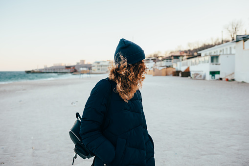 Unrecognizable young woman dressed in warm down jacket and hat stands with her hands in pockets looking away on winter sea sandy beach near water.