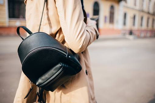 Close-up of elegant women's backpack. Rear view of young woman dressed in beige coat and with black bag walking down street in city.