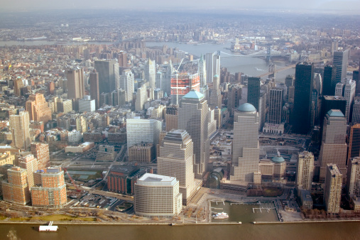 An aerial view of the Lower Manhattan skyline with the Hudson River in the foreground and the East River in the background. There are many tall skyscrapers, but also some lower level buildings. 
