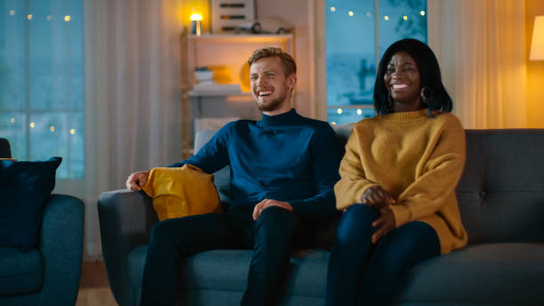 istock Happy Diverse Young Couple Watching Comedy on TV while Sitting on a Couch, they Laugh and Enjoy Show. Handsome Caucasian Boy and Black Girl in Love Spending Time Together in the Cozy Apartment. 1163710892