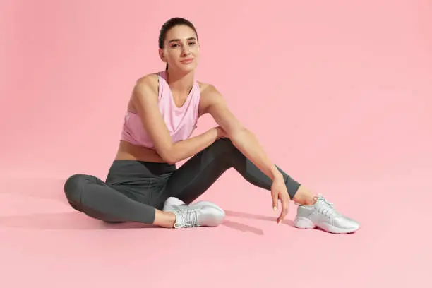 Fitness woman model in fashion sports wear relaxing on colorful pink background. Full lenght portrait of sexy girl with fit body in stylish sport clothes sitting in studio