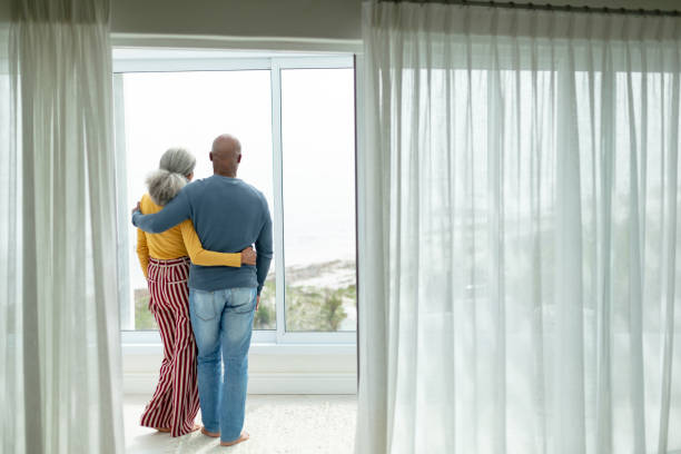 aktives seniorenpaar mit arm umstehend in der nähe des fensters zu hause - full length windows stock-fotos und bilder