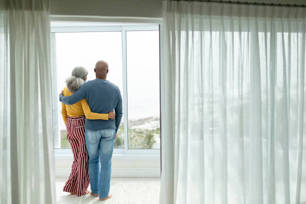 pareja activa de la tercera edad con el brazo alrededor de pie cerca de la ventana en casa - full length windows fotografías e imágenes de stock