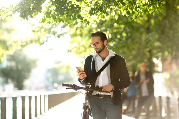 Businessman using smart phone while standing next to his electric bicycle Photo series of a young adult male commuting with electric bicycle through Berlin and doing business at the same time, drinking coffee from reusable coffee cup, listening to podcasts and making business calls. car city urban scene commuter stock pictures, royalty-free photos & images