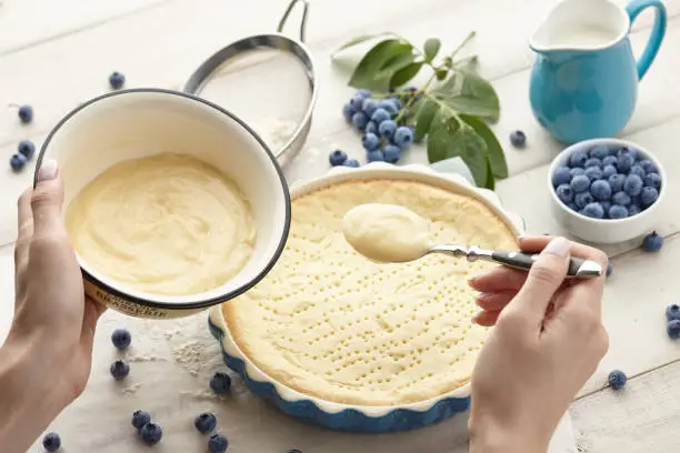 Woman pouring whipped cream into pie crust. Blueberry pie or tart cooking recipe.