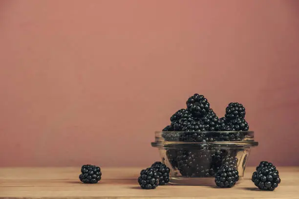 Fresh blackberry in glass bowl on a brown wooden table and dark-red wall background.