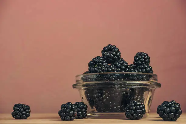 Fresh blackberry in glass bowl on a brown wooden table and dark-red wall background.