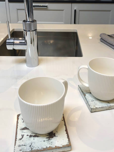 istock Image of modern kitchen design with central island breakfast bar / kitchen table with white composite corian worktop countertop and coffee mug / distressed painted square wooden coasters set for afternoon tea, cabinets, sink and mixer tap background 1163739641