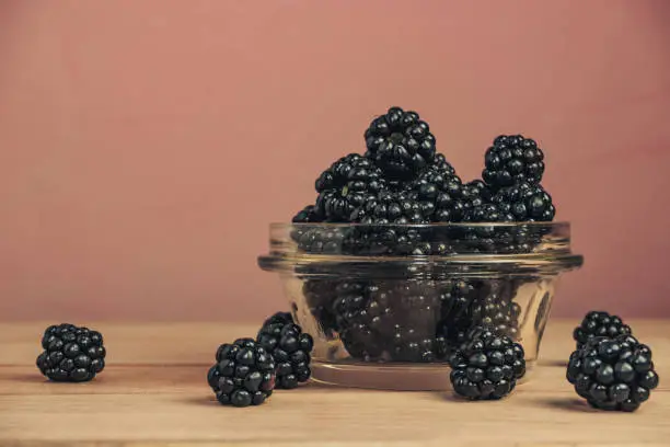 Fresh blackberry in glass bowl on a brown wooden table and dark-red wall background.