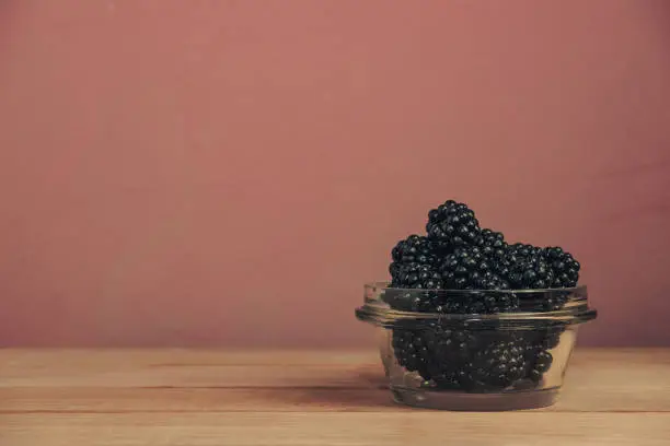 Fresh blackberry in glass bowl on a brown wooden table and dark-red wall background.