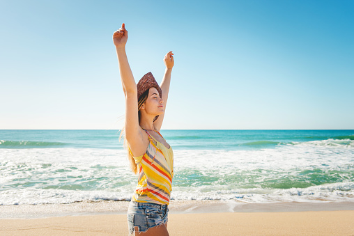 Young woman in straw hat with arms raised enjoying the breeze on the beach in a hot summer day