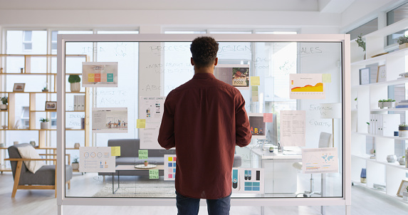 Rearview shot of a young businessman writing down ideas on a glass wall in his office