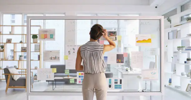 Rearview shot of a young businesswoman scratching her head while writing down ideas on a glass wall in her office